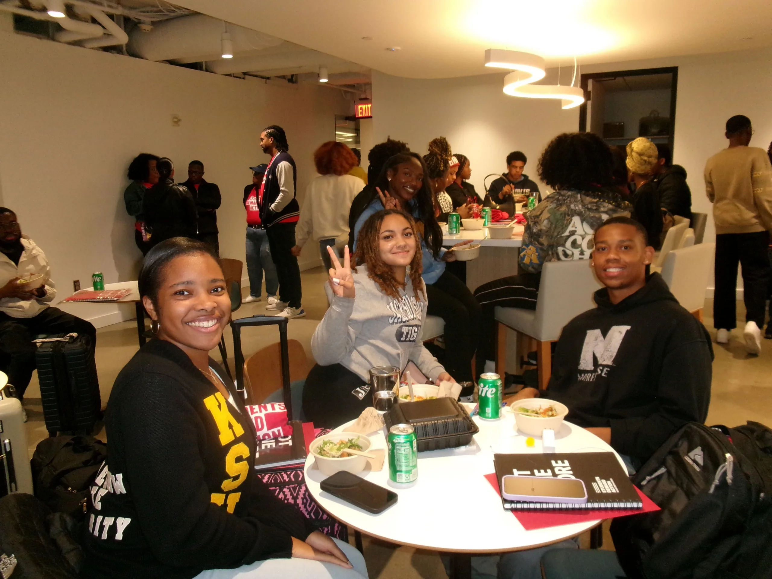 Three students pose for a photo at the Young Changemakers Summit. Approximately 20 other people are visible in the background.