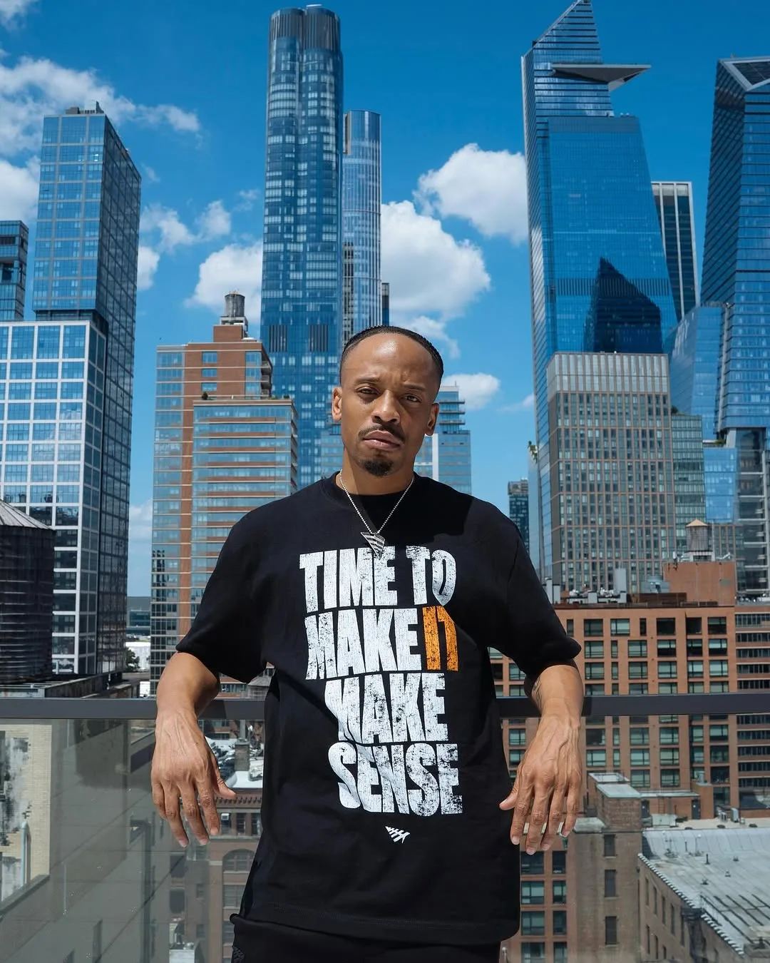 Justin Murray wears a black t-shirt with white block letters that read "Time to Make It Make Sense" with the Paper Planes logo beneath the words. Justin stands on a balcony with a glass wall in front of an NYC skyline with blue sky and white clouds behind him.