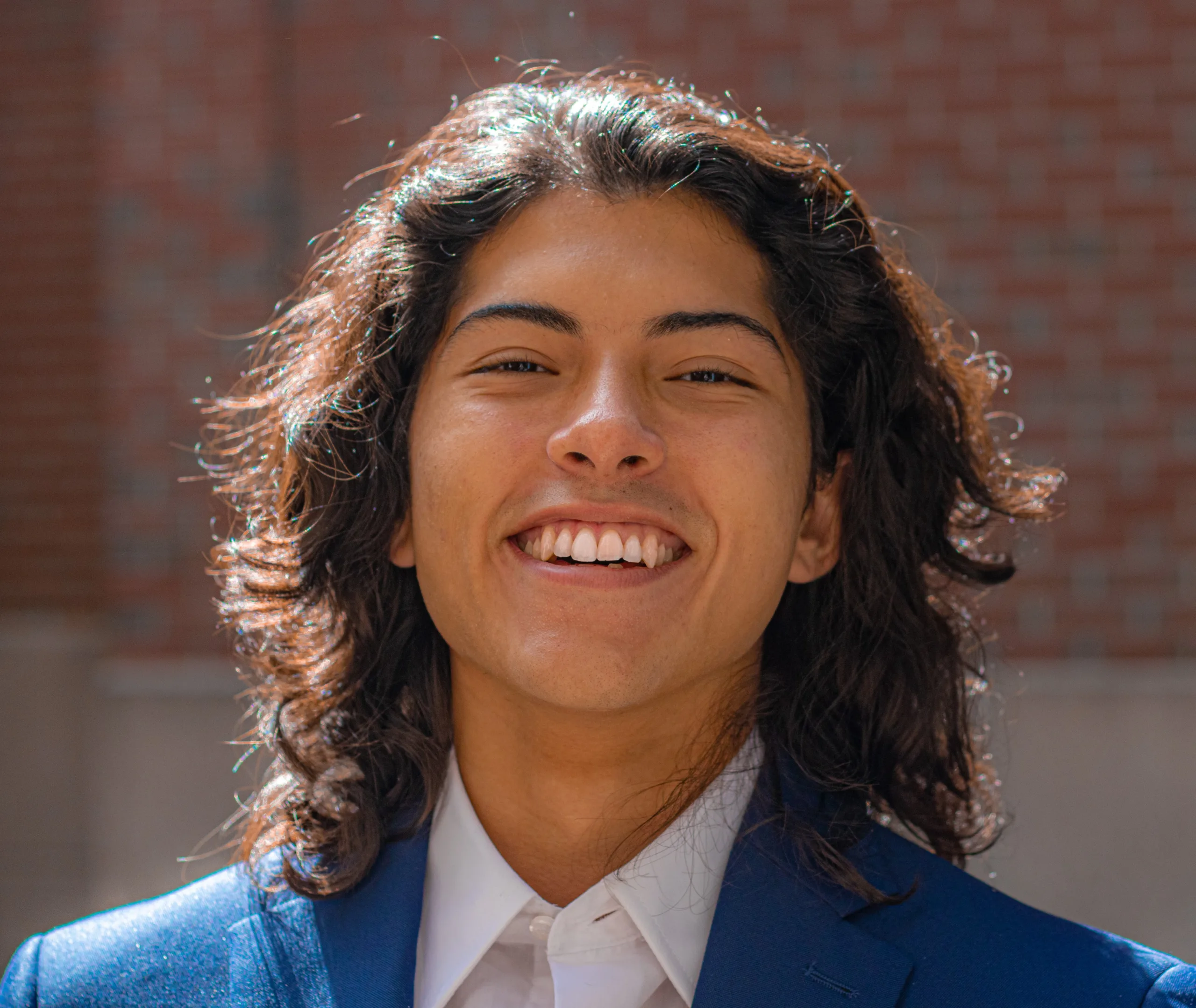 Andres Cubillos is pictured in a shoulder-up headshot. He is wearing a blue suit jacket and a white collared shirt with the top button unbuttoned. He has dark curly hair that is cut to his shoulders and parted in the middle.