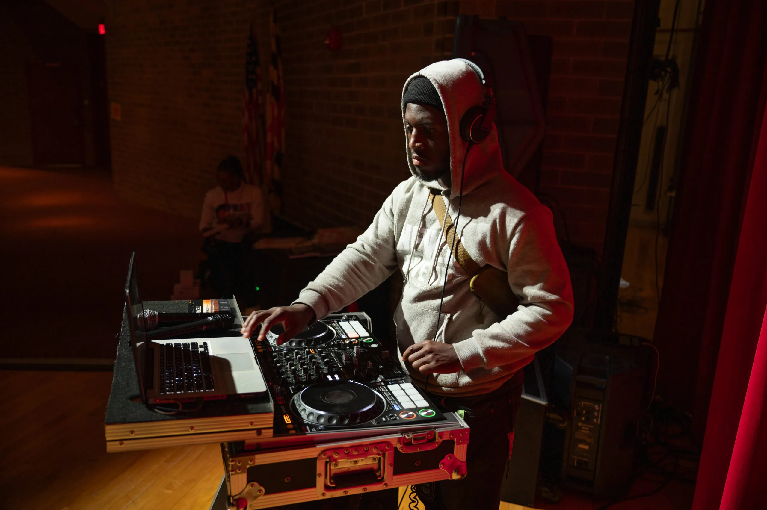 A DMV-area DJ stands under stage lights while playing music from a turntable and laptop. The DJ wears a light hoodie, over-ear headphones, and a black beanie.