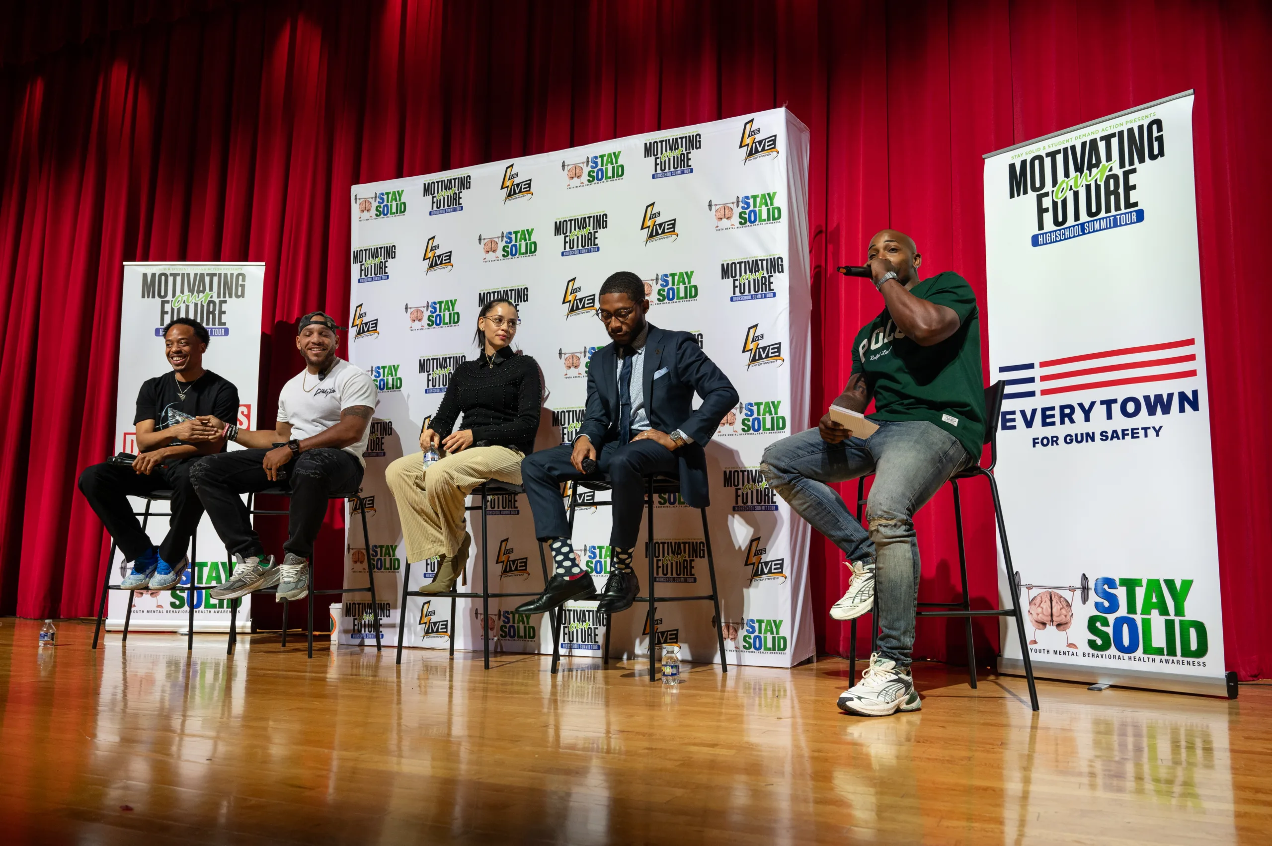 Neal McDonald (far right) wears a hunter green t-shirt and jeans as he speaks to an off-camera crowd at a Motivating Our Future tour stop. Four other panelists smile and sit beside him on stage as they listen.