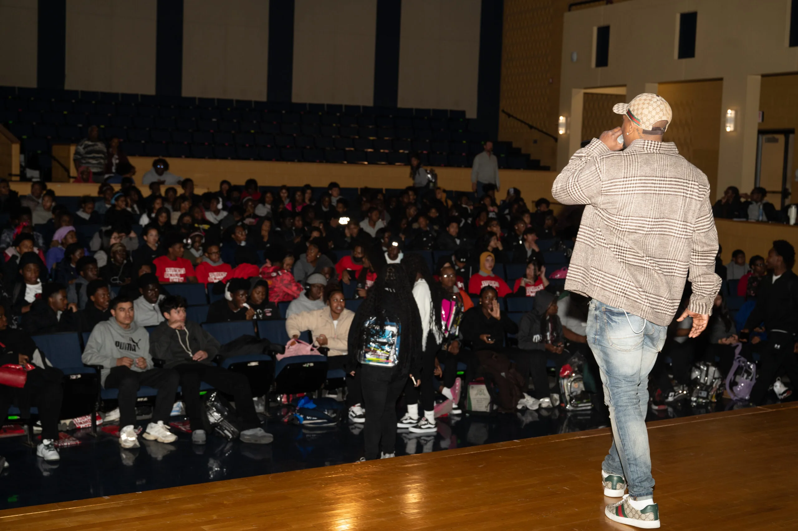 Neal McDonald is pictured from the back as he speaks from a stage out to a crowd of students in a high school auditorium. Many students are wearing red Students Demand Action t-shirts. Neal wears a brown striped flannel, a checkered baseball cap, and light-wash jeans.