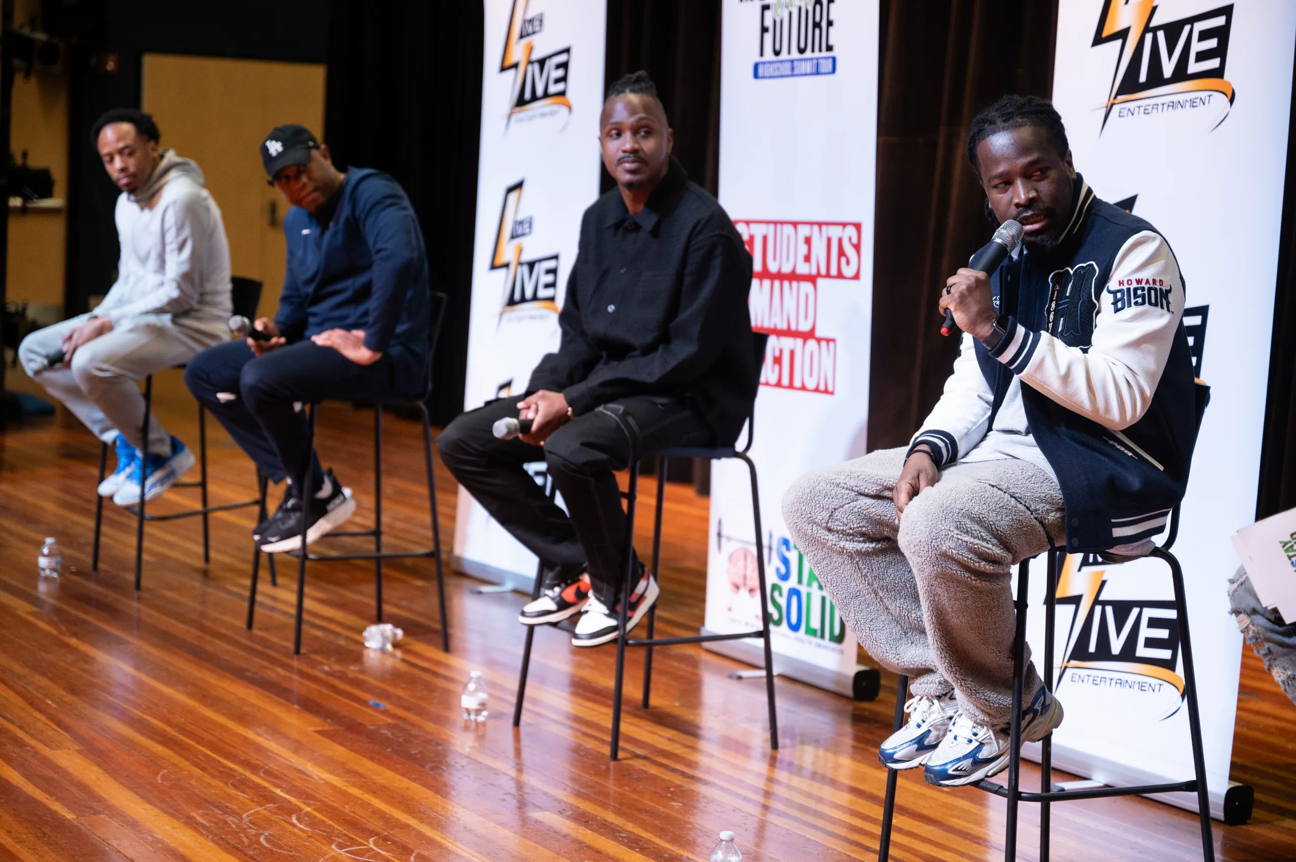 Panelists at the Motivating Our Future Summit Tour look to their left at a fourth panelist, who is holding a microphone and looking off camera as he speaks during the Motivating Our Future Summit Tour.