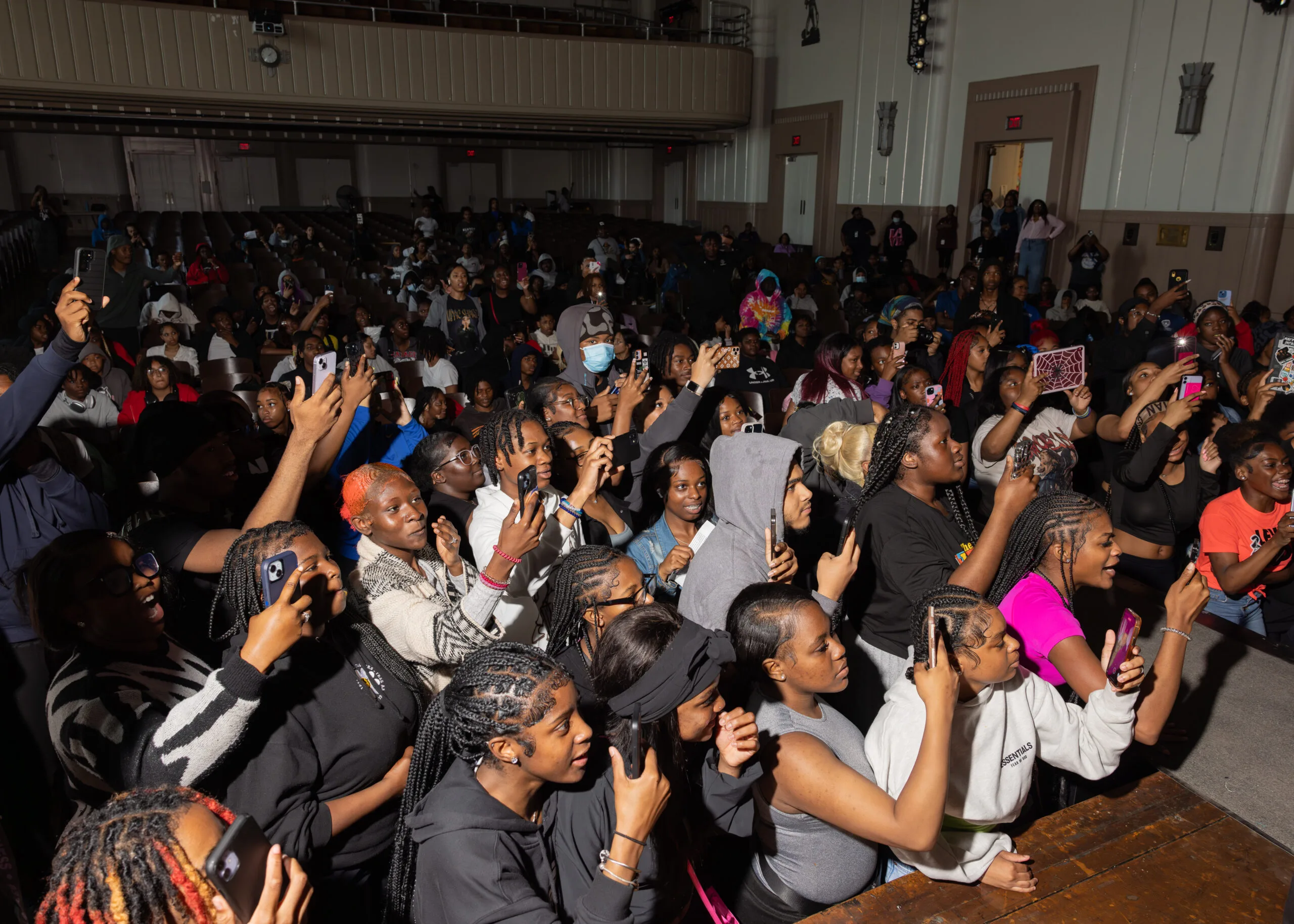 Black students attending the conflict resolution tour gather toward the stage; many of them have their phones or other recording devices out and are pointing the cameras toward the stage, filming a performance.