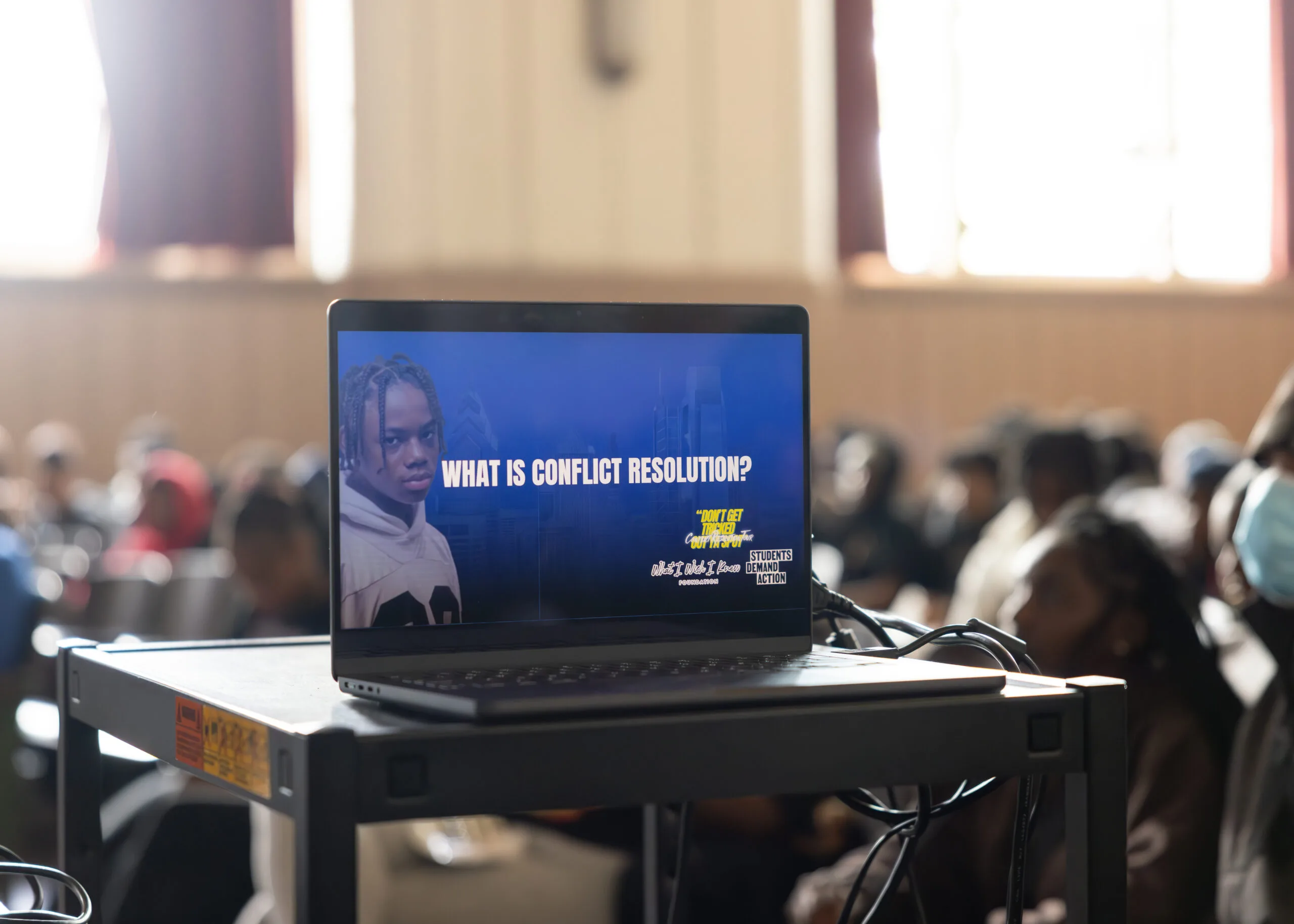 A photo of a laptop, set on a rolling projector cart; the laptop is in focus, while the students in the background behind it are blurred. The slide has a blue background with a Black student pictured on the far left; the student has braids and a white sweatshirt. In white text centered on the slide, the words "What is conflict resolution?" appear in all caps.