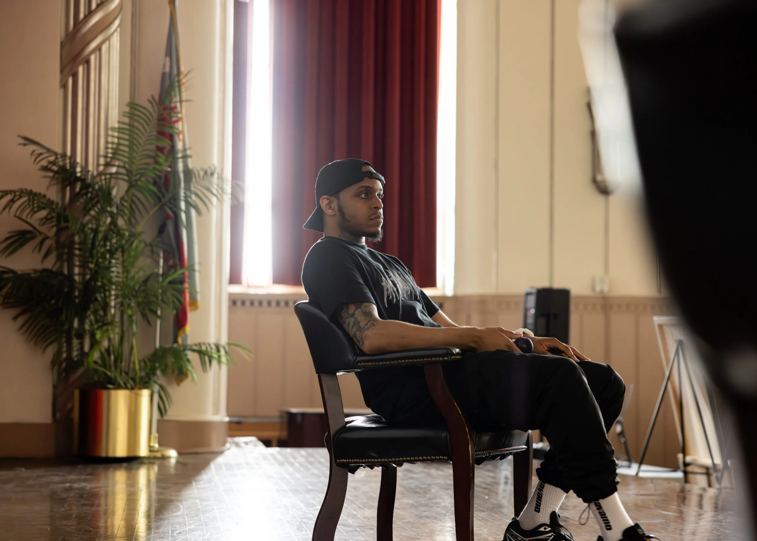 A photo of Manny taken at a side profile. Manny is sitting on a black leather chair with gold studs and brown legs. Manny wears a black baseball cap flipped backward, a black t-shirt, black pants, white socks, and black sneakers. Behind his chair, you can see (from left to right) a green plant in a gold pot, a flag, and a red velvet curtain.