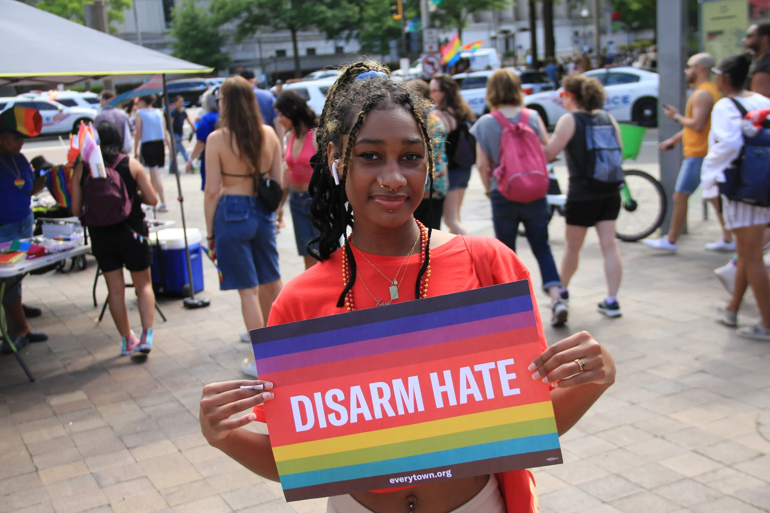 A Black student holds a rainbow sign that reads "Disarm Hate" in white letters. They wear their black hair in braids gathered into a ponytail; two braids frame their face. The student has a white AirPod in their right ear.