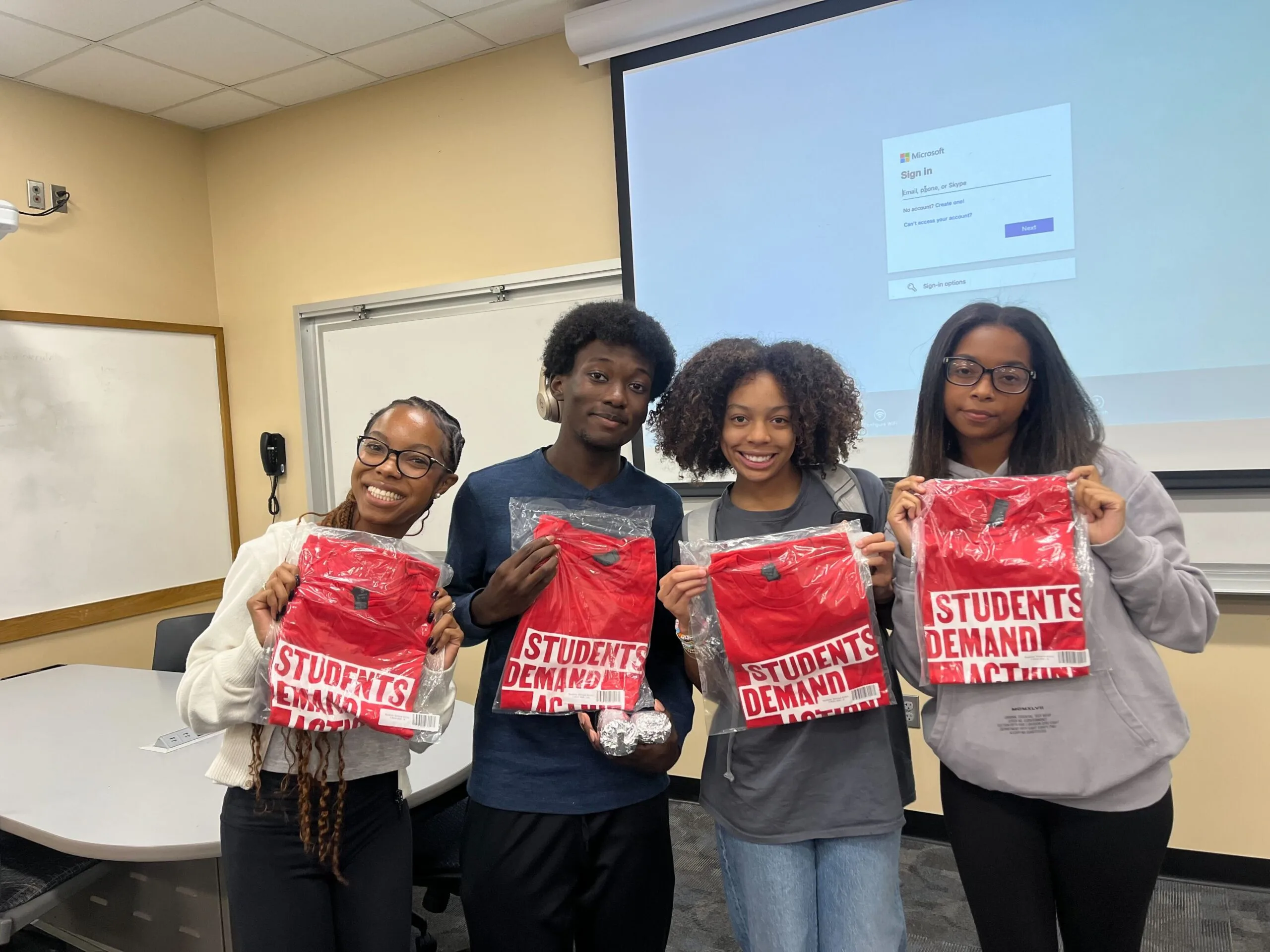 A group of four Black students stand in a row and smile at the camera; they are holding new Students Demand Action red t-shirts that are in new plastic packaging. A projector screen and two whiteboards are visible in the background.