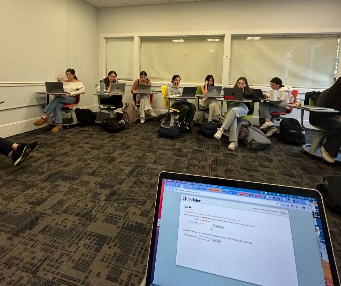 A group of around 10 students phonebank in a classroom. The students are sitting at desks and have their laptops open; they are holding their phones up to their ears. In the foreground of the photo, a laptop screen with the Hubdialer log-in screen is visible.