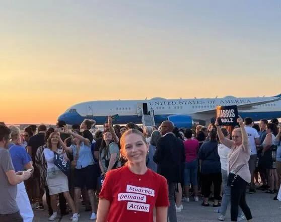 A student in a red t-shirt that says "Students Demand Action" poses for a photo in front of a crowd of people, who are gathered on the tarmac to greet Harris and Walz as they arrive to a rally. The sky is a gradient from orange near the horizon to blue near the top of the image frame and appears to be sunset. A plane is visible in the background; it says "United States of America" in all-capitals text.