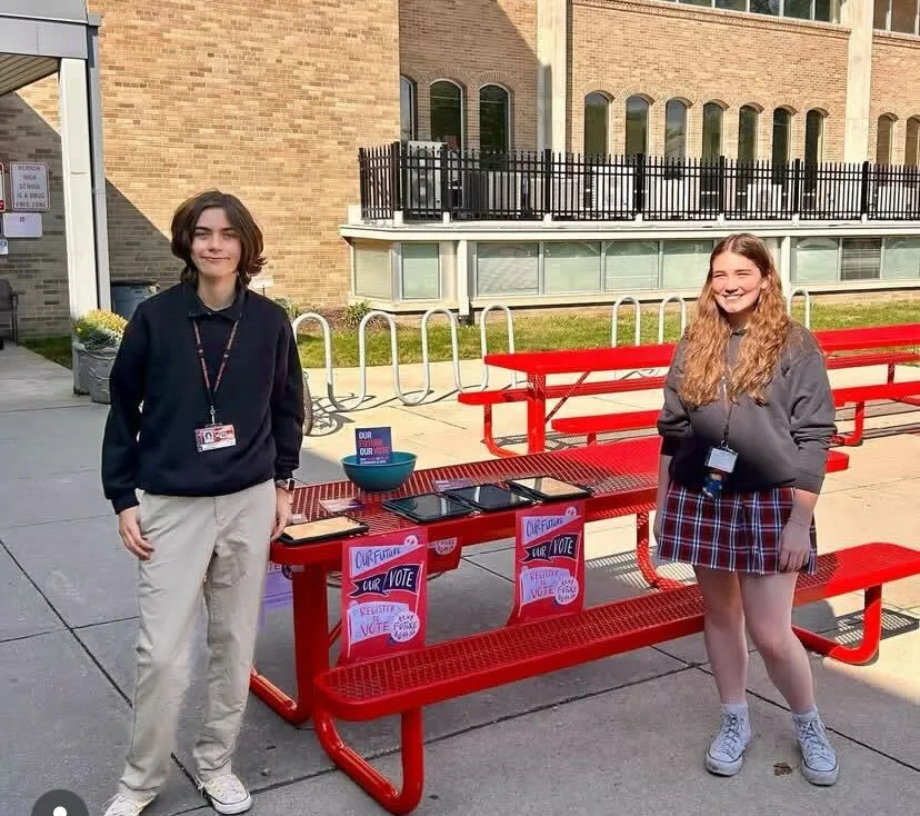 Two Students Demand Action volunteers stand in front of a red metal picnic table. The table has several tablets sitting on it to help students register to vote; several posters are propped on the table's bench seat that read "Our future. Our vote. Register to vote. Text Future to 644-33."
