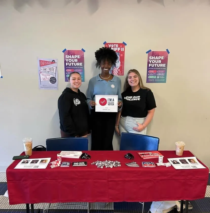 Three Students Demand Action volunteers at Drake University pose for a photo while tabling. They stand behind a table with a red tablecloth that has stickers, candy, and literature about Students Demand Action. One student wears a black t-shirt that says "end gun violence." Another holds a sign that reads "I'm a Gun Sense Voter."