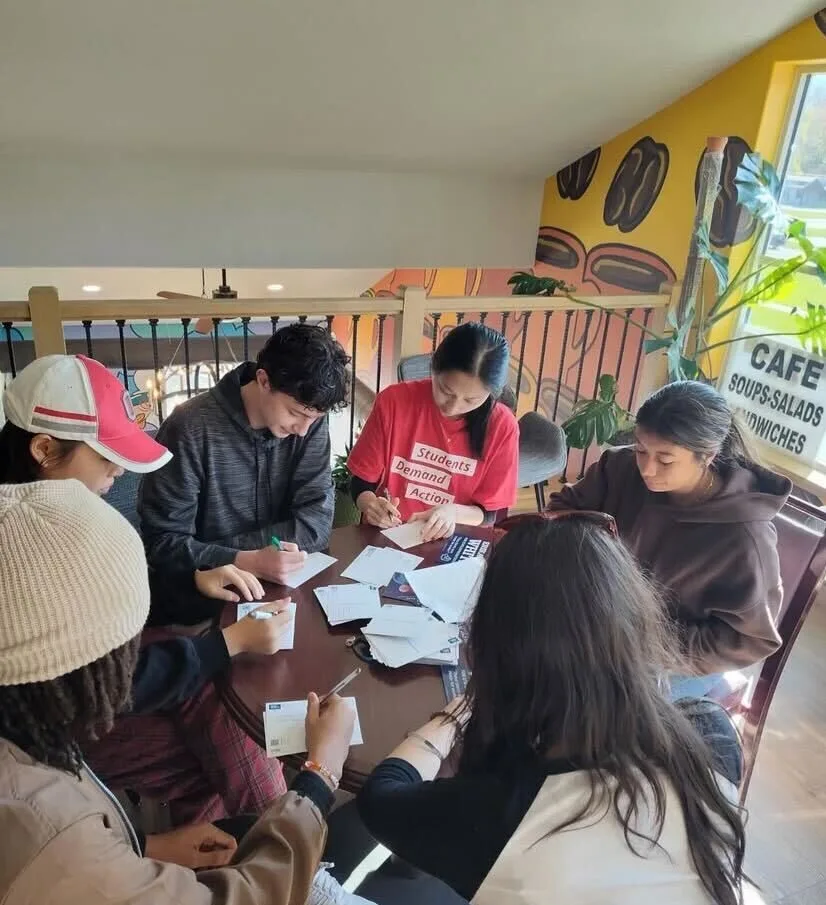 A group of six Students Demand Action volunteers sit at a round table in a cafe. They are looking down at the table while writing on white postcards.