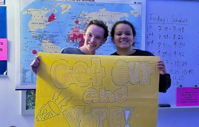 Two students stand in front of whiteboard in a classroom that has a map of the world pinned onto it and "Friday's schedule" written in black marker. The students are holding a handmade yellow poster that says "Get Out and Vote!" in block letters.