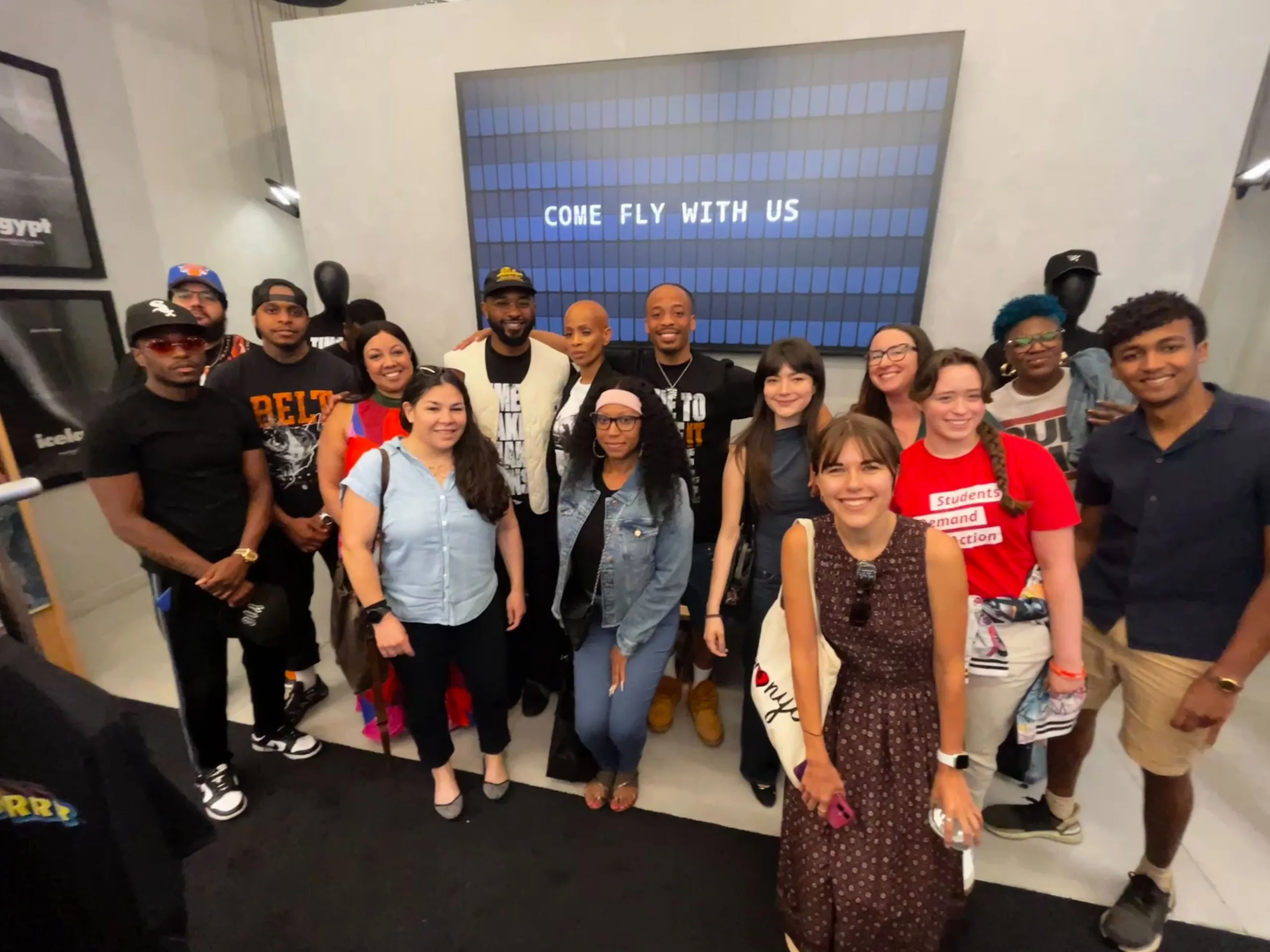 Justin Murray, Neal McDonald's mother, and 14 other people pose for a photo during the Paper Planes pop-up event in memory of Neal McDonald. Behind them, a blue grid screen reads "Come Fly With Us" in white letters.