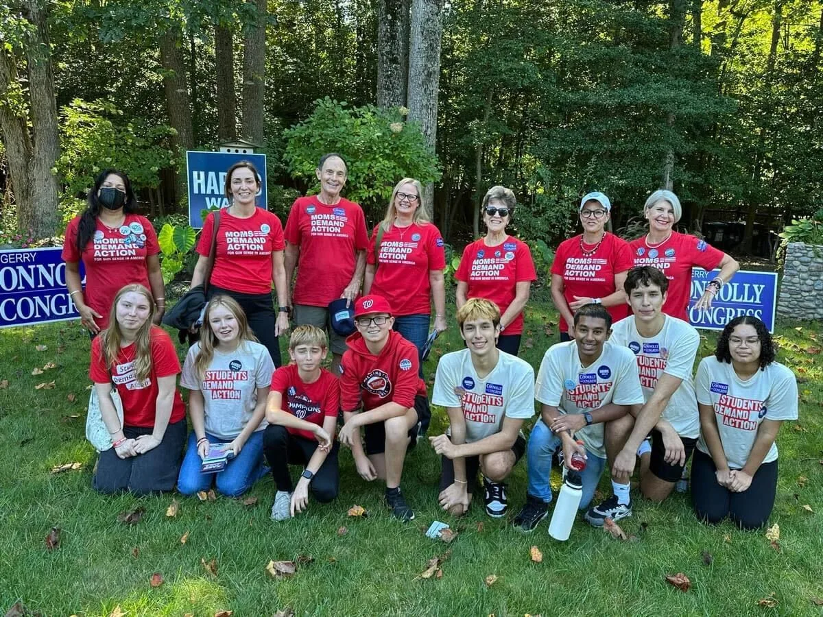 A group of 15 total Moms Demand Action and Students Demand Action volunteers pose for a photo. In the front row, 8 students kneel; in the back row 7 adults stand.