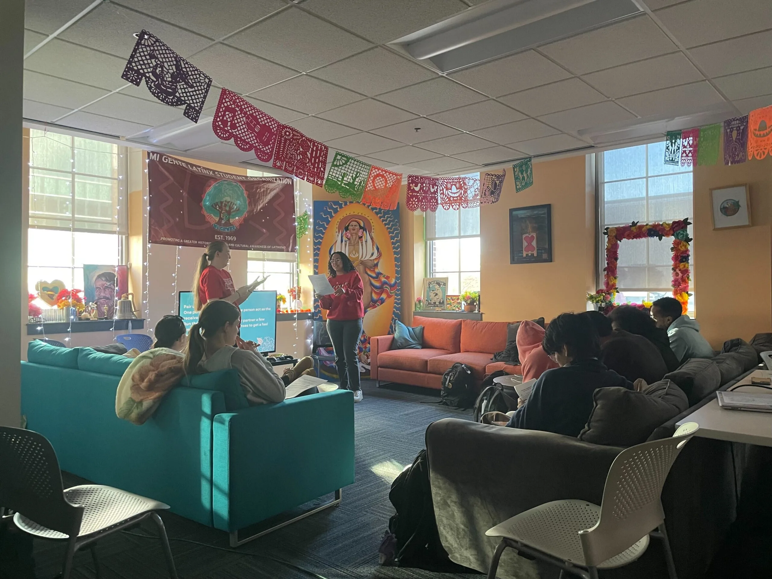 A candid photograph of Students Demand Action and Mi Gente group members at the University of Minnesota. Students are sitting on teal, orange, and grey sofas while watching two Students Demand Action volunteers act out how to use a phonebanking script. Papel picados in shades of purple, orange, red, and green hang from the ceiling.