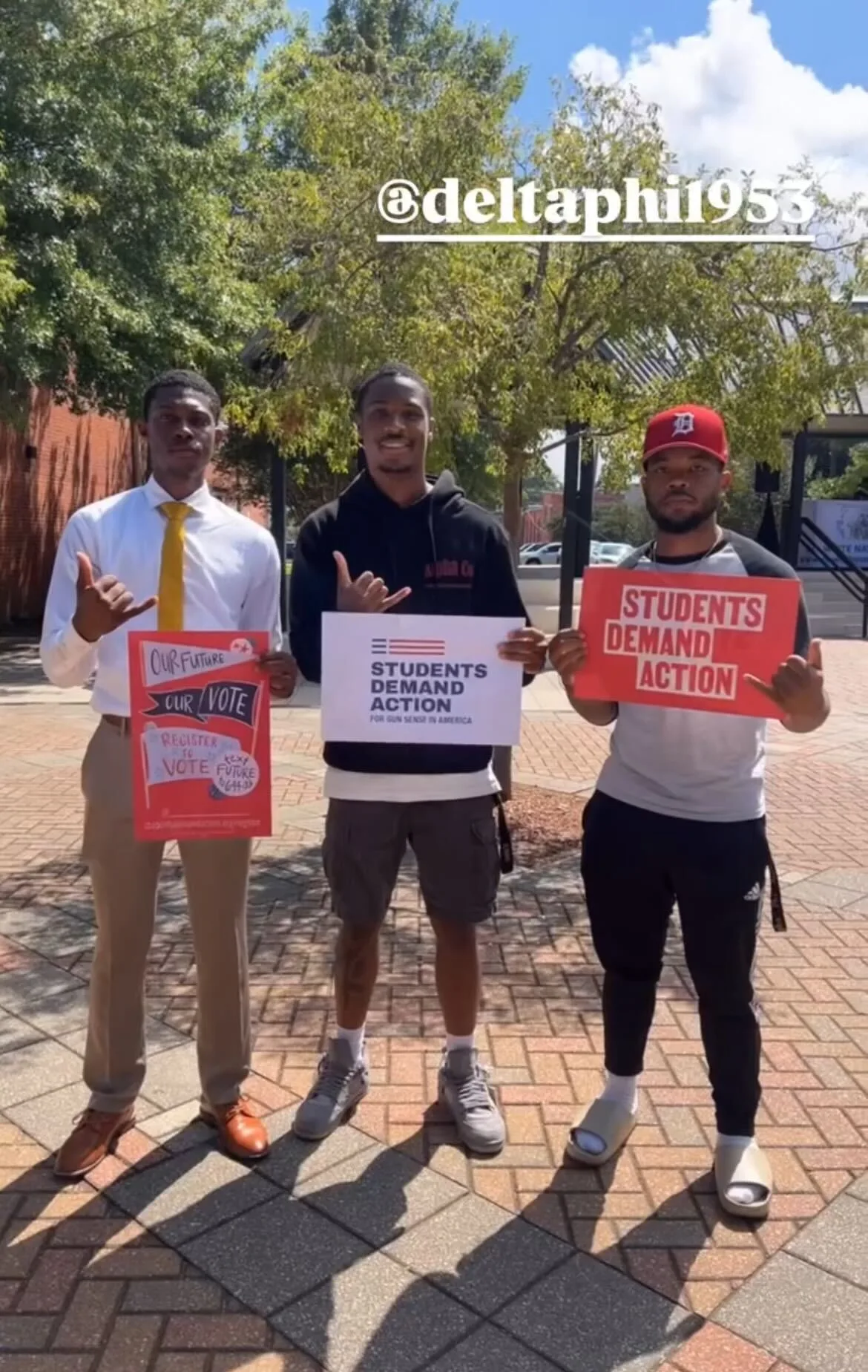 A group of three Black men hold Students Demand Action signs and pose for a photo on a campus quad. One sign reads "Our future. Our vote. Register to vote. Text "Future" to 644-33." The other two students hold signs that say "Students Demand Action." The photo has an Instagram handle tag @deltaphi1953.