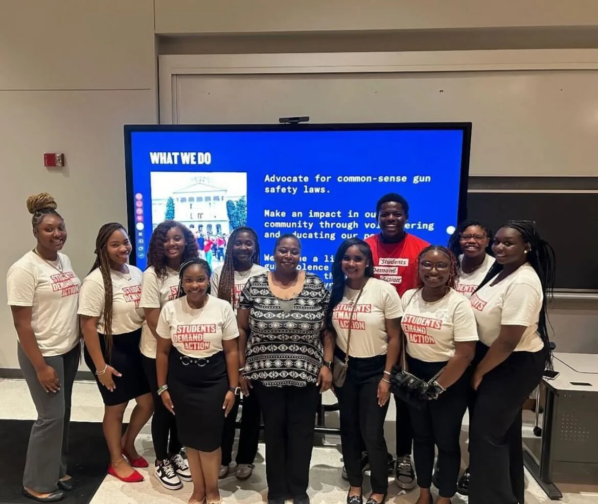 A group of 10 Black students pose for a photo in front of a blue projector screen displaying a slide titled "What We Do." The students are wearing white Students Demand Action t-shirts with a red block-text logo. One student in the back right of the group wears a red Students Demand Action t-shirt.