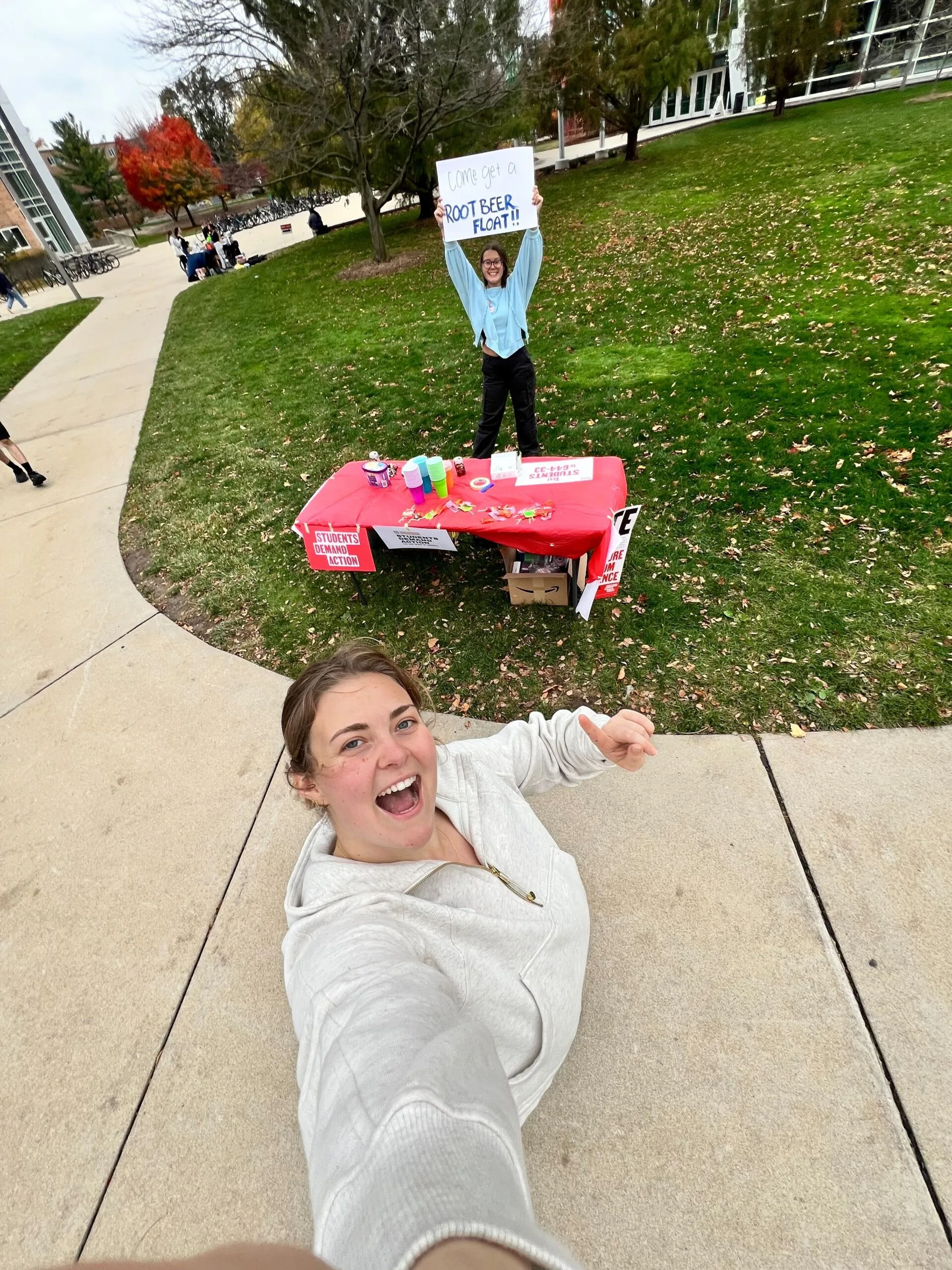 Two Students Demand Action volunteers take a selfie while tabling on their campus quad. One of them holds a hand-written sign that reads "Come get a ROOT BEER FLOAT!!"