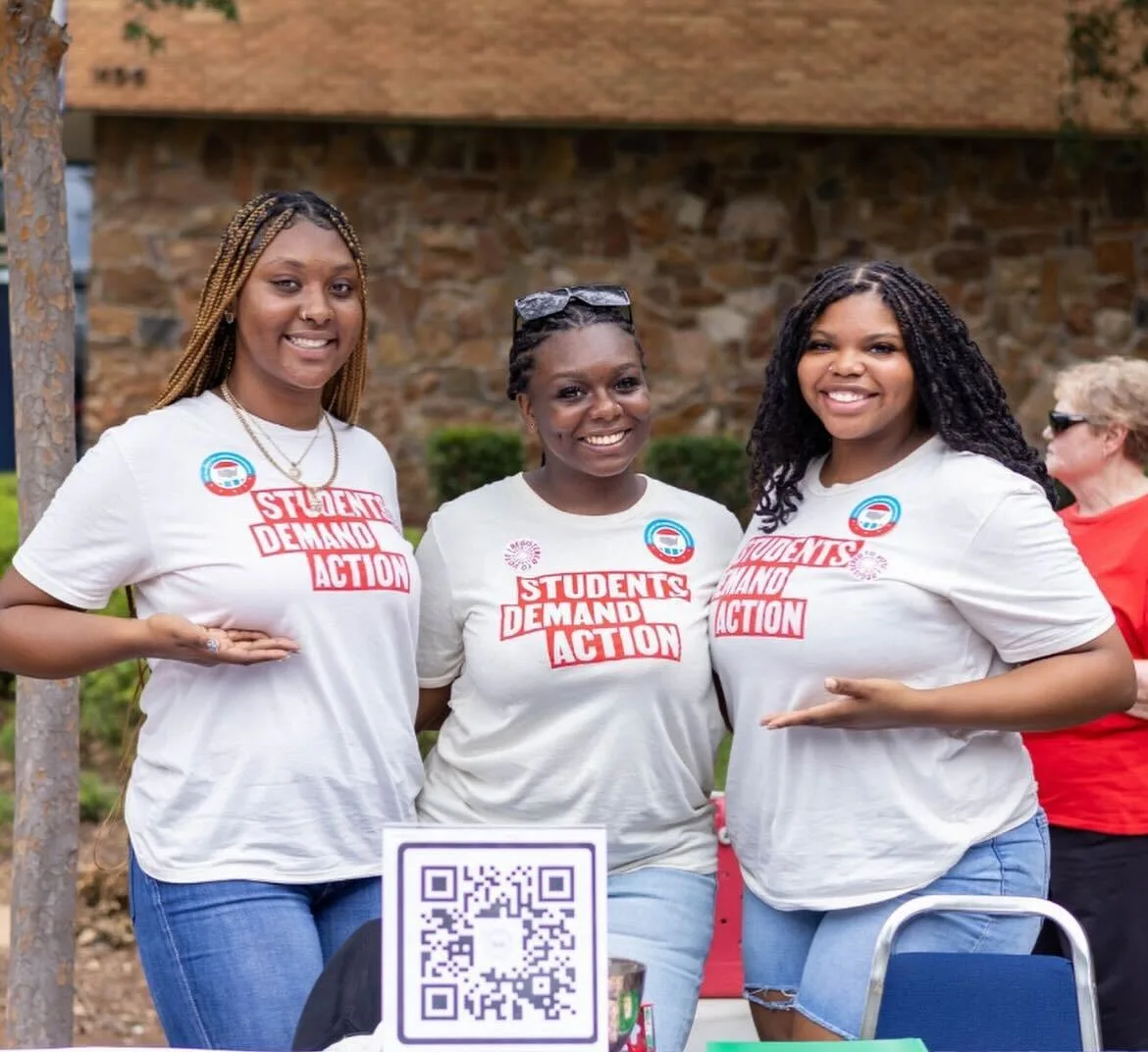 Three Black students wearing white Students Demand Action t-shirts with red block lettering post for a photo in front of a QR code to register to vote while tabling on their campus plaza. A white woman in a red t-shirt and dark sunglasses is visible in the background on the far right of the photo.