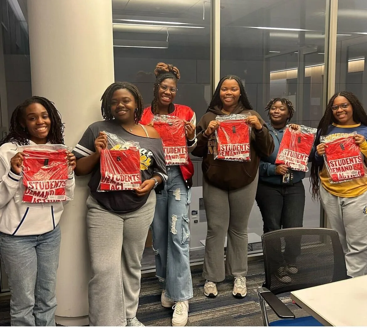 Six Black women stand in a row holding new Students Demand Action red t-shirts; the shirts are in clear plastic packaging.