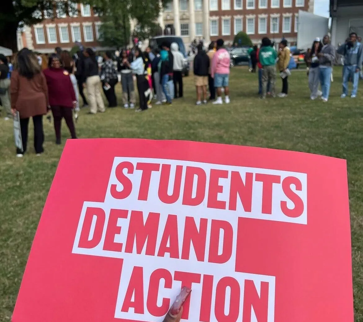A red Students Demand Action sign is in focus in the foreground. In the background, students stand in a half-circle in a green space on campus.