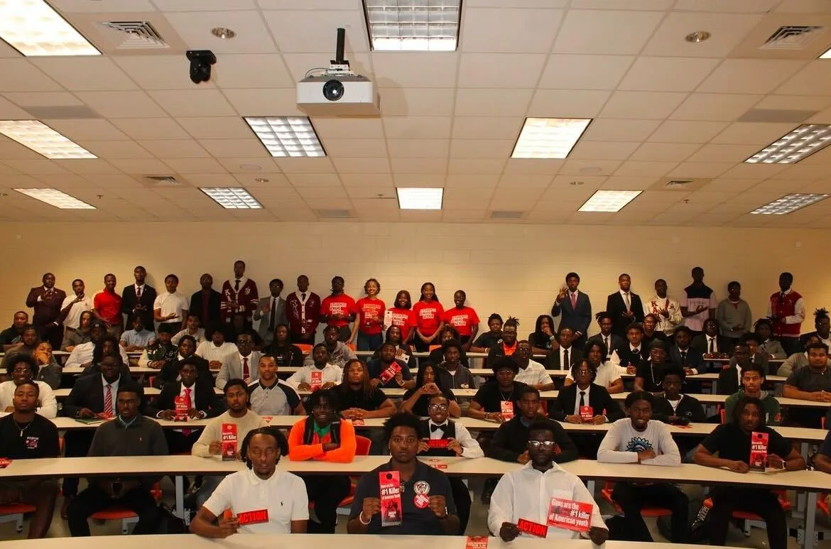 A room full of Black students at Florida A&M University, an HBCU, sit in 6 tiered rows of long desks in a large classroom on campus. A seventh row of students stands behind the last row of desks.