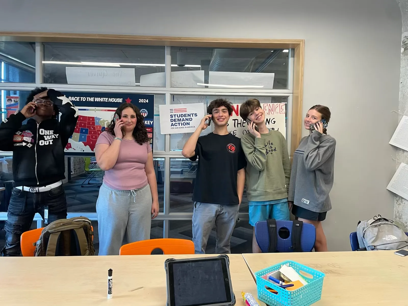 Five Students Demand Action volunteers at the University of Florida pose for a photo. They stand behind two light-colored wood tables with plastic orange and blue chairs. Each student is holding a cell phone up to their ear.
