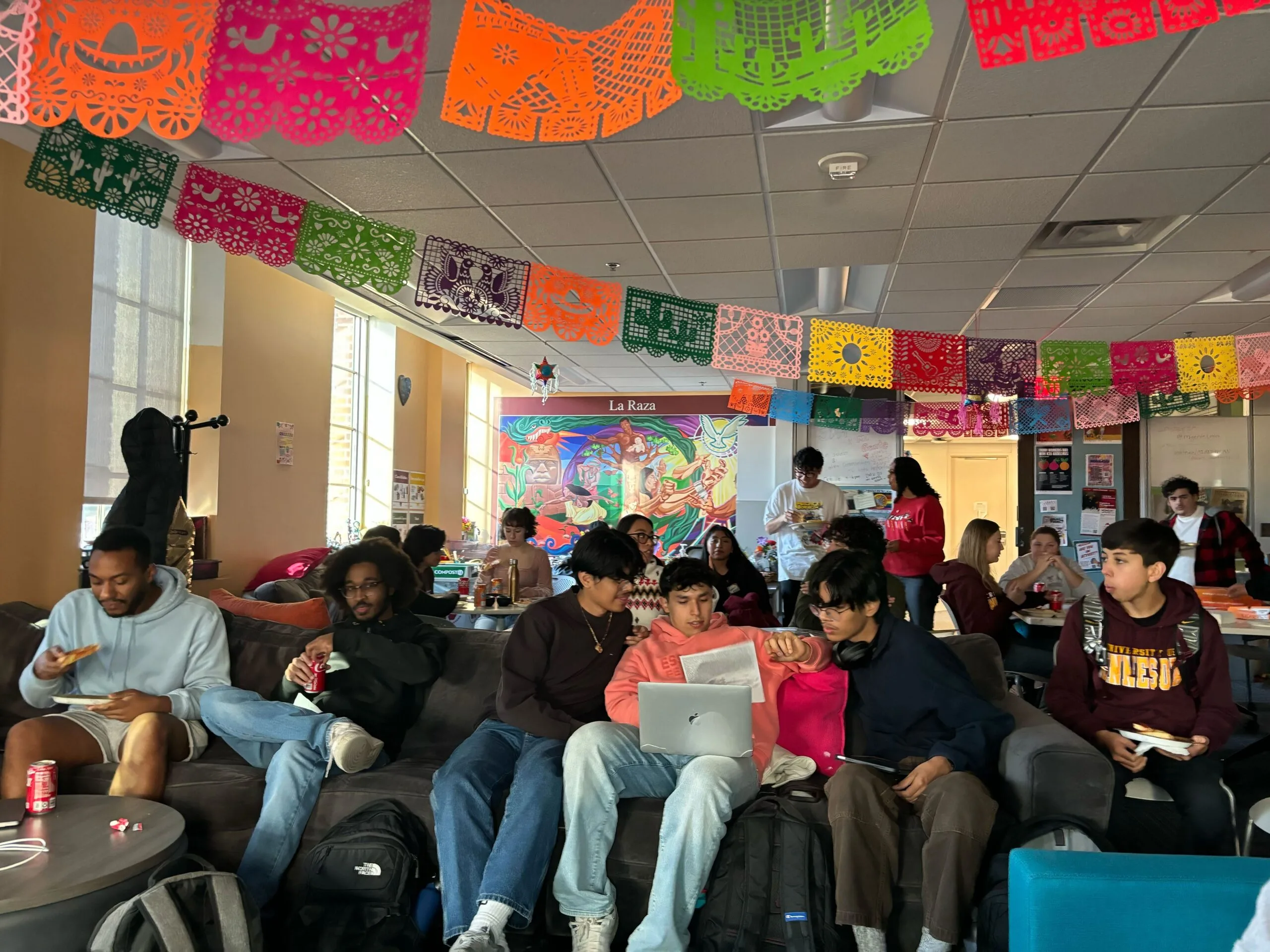 A group of approximately 20 University of Minnesota students are pictured in a candid photograph. Papel picados in orange, pink, green, yellow, and blue shades hang from the ceiling. Students are sitting on couches and at tables, eating pizza and learning how to phonebank.