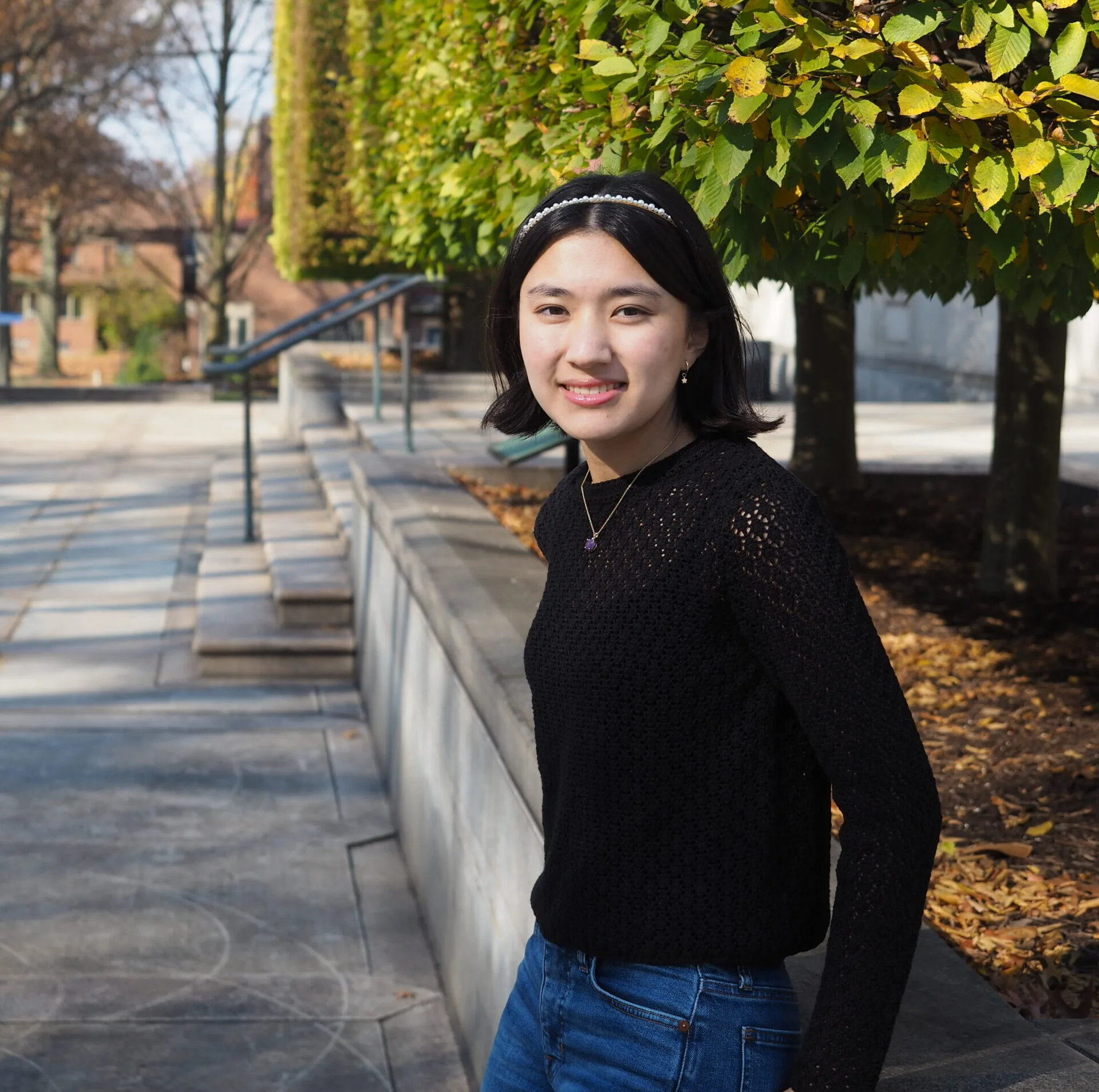 Pearl Lee, a Students Demand Action Text Team Captain, poses for a photo outside. Pearl wears a black lace-knit sweater, a pearl headband, and blue jeans.