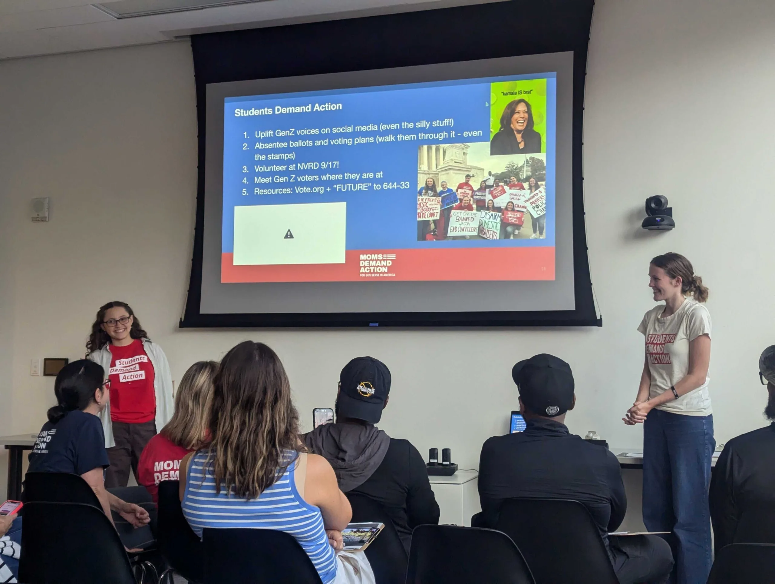 Two Students Demand Action volunteers present to a group of Moms Demand Action volunteers. The students stand on each side of a presentation screen. The slide includes 5 ways that Moms Demand Action volunteers can include Students in their elections work. The top right of the slide has a meme with a picture of Kamala Harris on a lime green background, captioned "kamala IS brat"