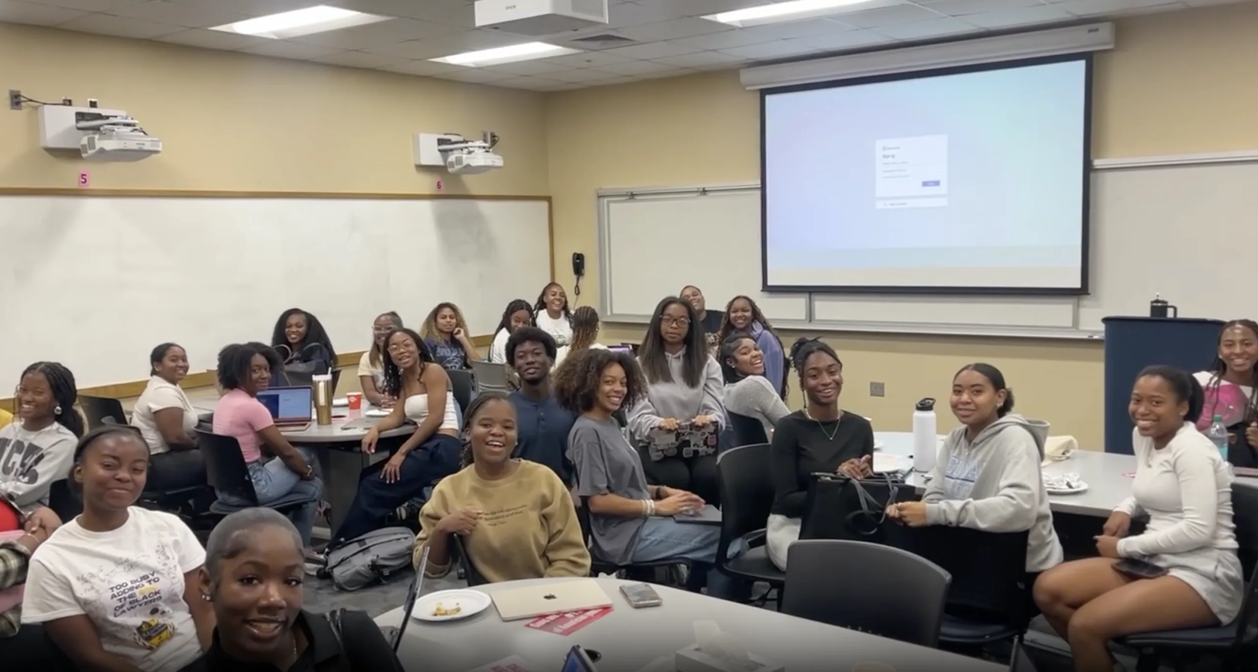 A group of around 25 Black students smile for the camera in a group photo in what appears to be a classroom. The students are sitting at various oblong tables around the room. A projector screen and two whiteboards are visible in the background.