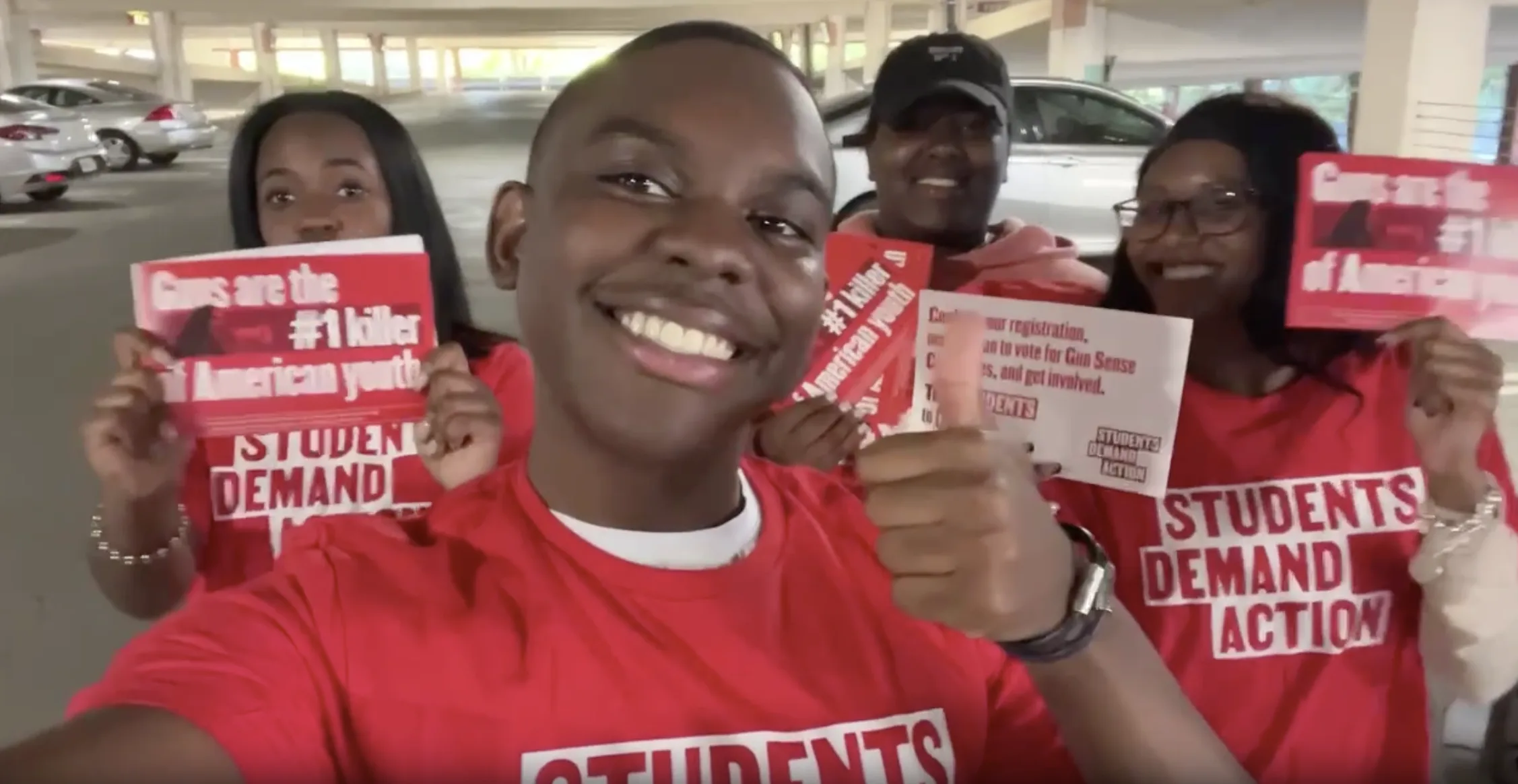A group of four Black students wearing red Students Demand Action t-shirts take a selfie together. Three of them hold cards reading "Guns are the #1 killer of American youth." The student taking the selfie is holding the phone with one hand and giving a thumbs-up with the other.