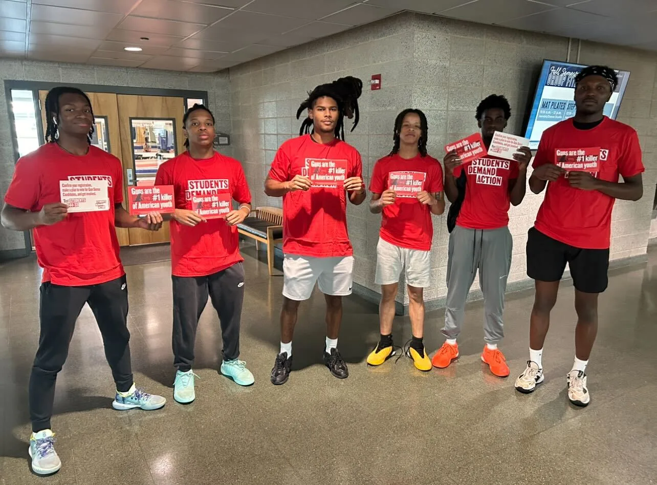 A group of six Black students from Georgia State University pose for a photo while vote tripling at their polling location. They all wear red Students Demand Action shirts and are holding red palm-cards that say "Guns are the #1 killer of American youth"