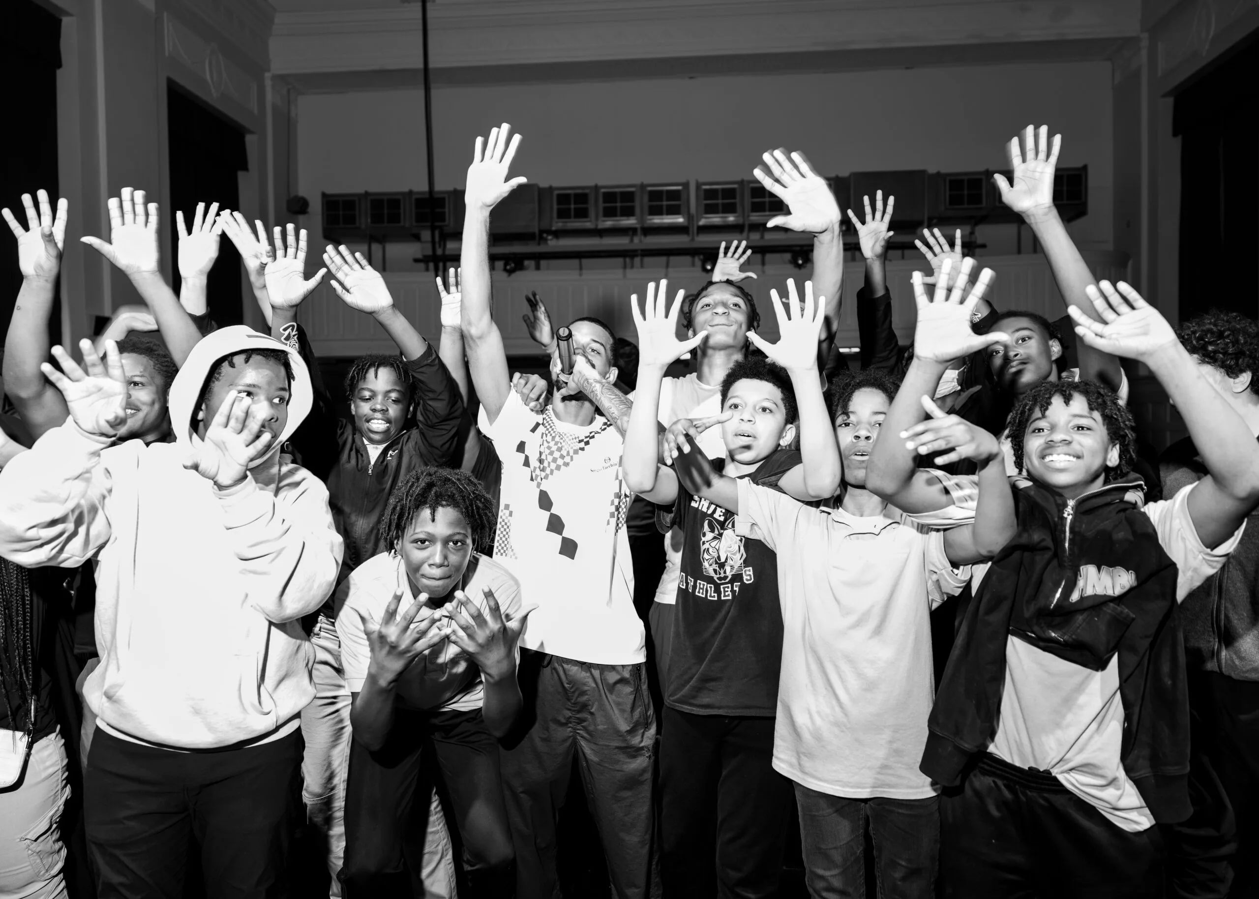 At least 12 students are pictured with a performing artist at one of the schools on the conflict resolution tour. The image is black and white, and all the students are holding both their arms up, waving both hands with their palms facing the camera. The artist has his right arm raised and is holding a microphone with his left.