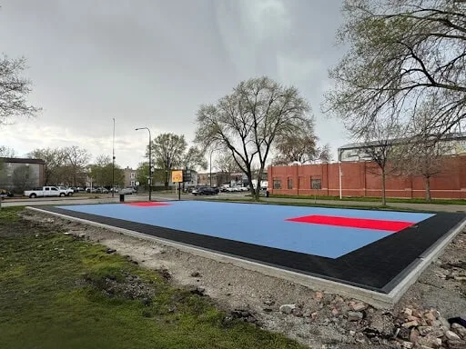A basketball court partway through the process of being renovated. The photo is taken at an angle from one corner; the court is light blue with bright red accents under the hoops; the border of the court is black. What appears to be a school building is visible in the background.
