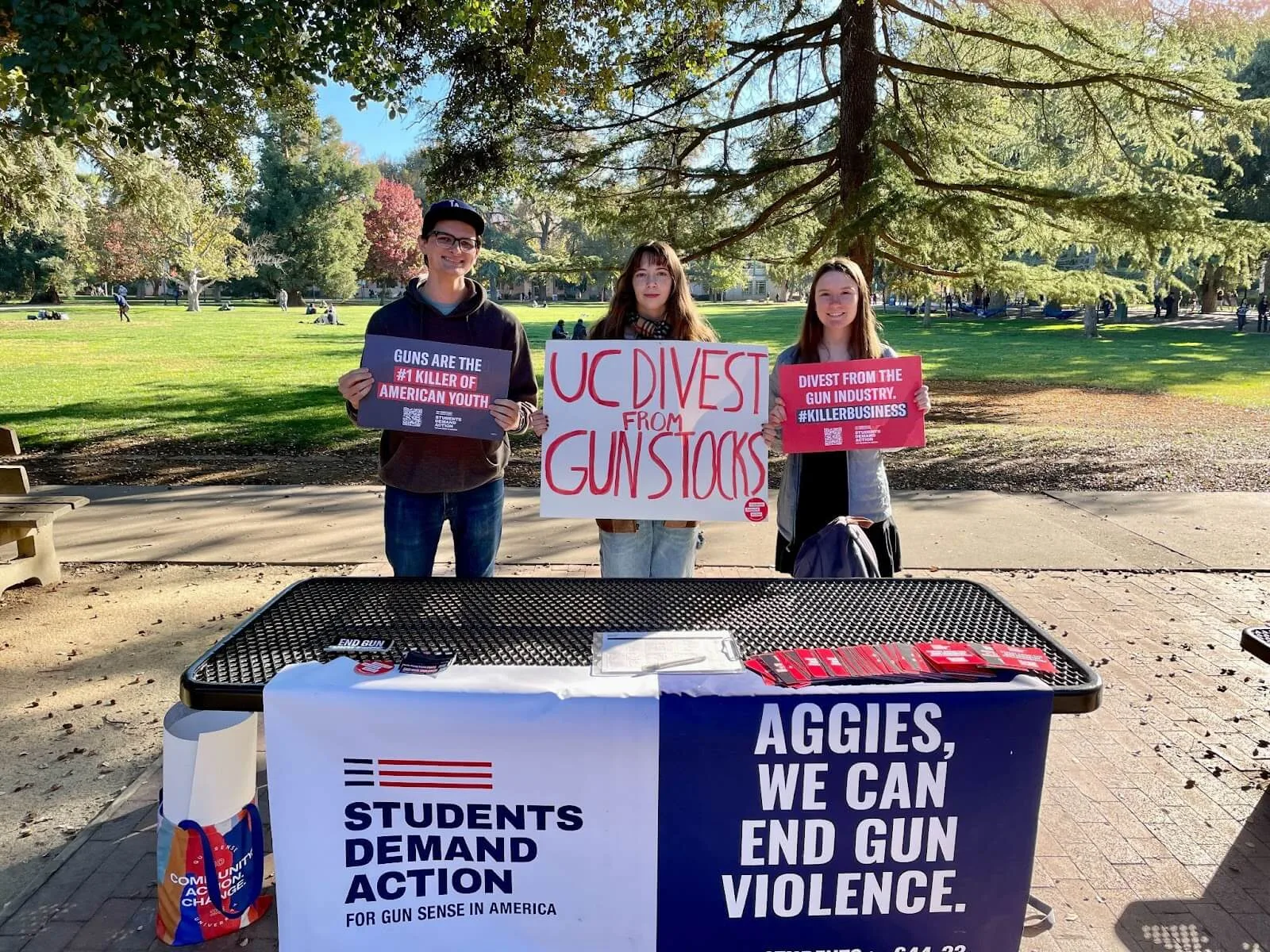 Three Students Demand Action volunteers table during divestment week of action. Each of the three people are holding signs. From left to right, they read: "Guns are the #1 killer of American youth"; "UC Divest From Gun Stocks"; "Divest from the gun industry. #KillerBusiness." On the front of the table, two large banners hang side-by-side; one is white with the Students Demand Action logo printed in large text. The other is navy and reads "Aggies, we can end gun violence."