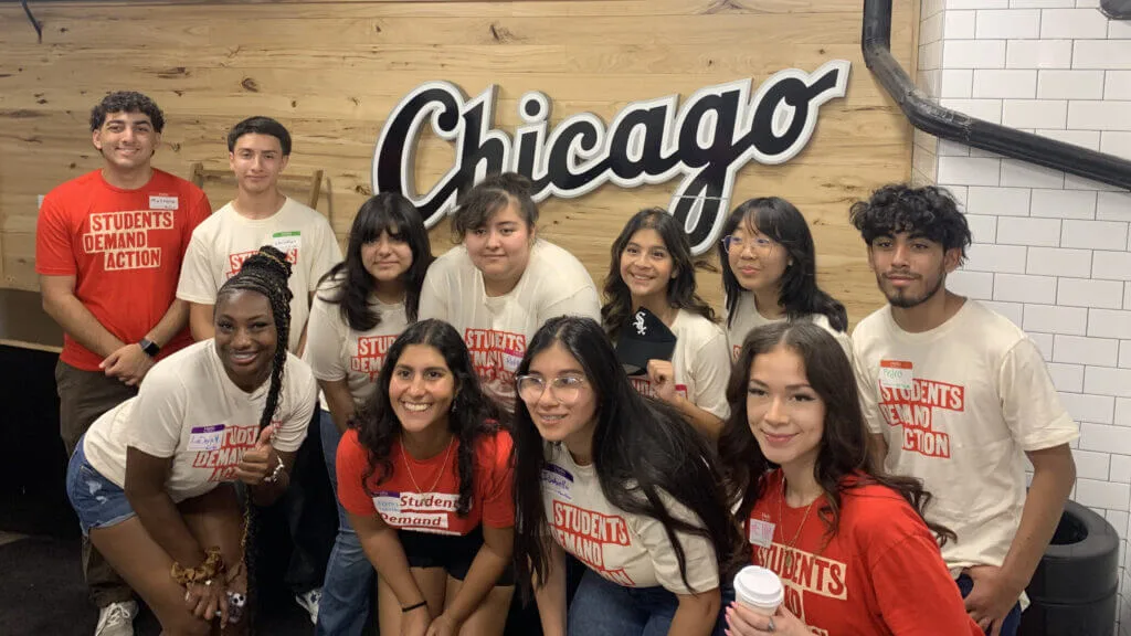 A group of students pose in front of a sign in the White Sox stadium that says "Chicago." They are all wearing red or cream-colored Students Demand Action t-shirts.