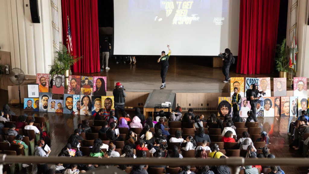 Manny, a Black man in his mid-20s, stands in the center of a brown stage in front of a projected slide that reads "Don't Get Tricked Out Ya Spot" Conflict Resolution Tour. Manny is raising his left hand and holding a mic to his mouth with his right. The photo is taken from a raised balcony; the backs of students attending the tour are visible in the photo.