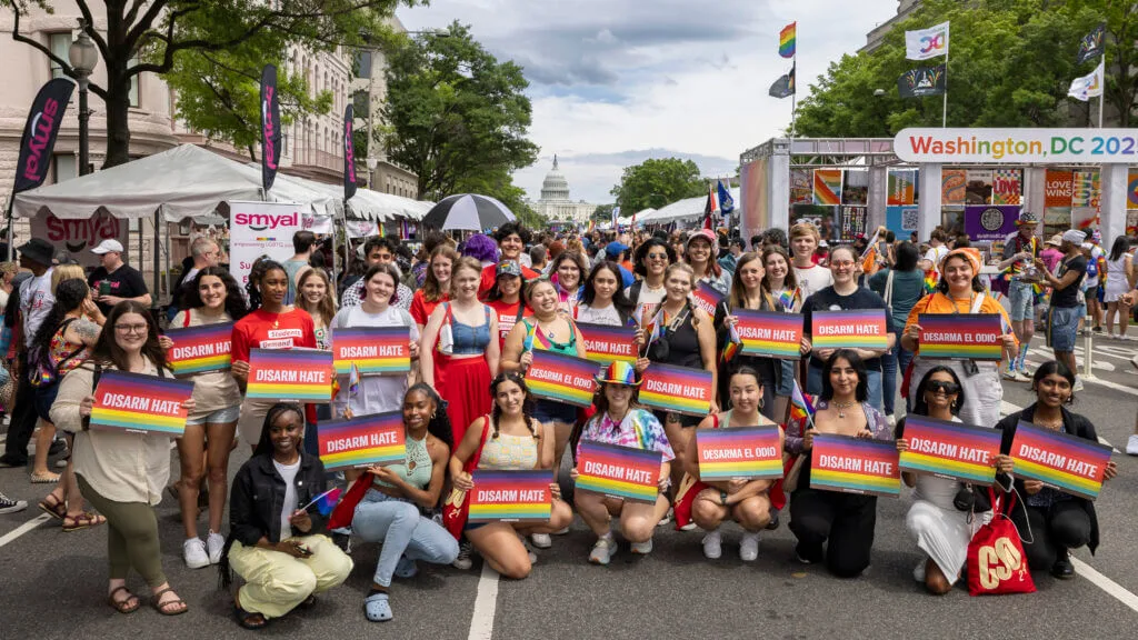 A group of about 20 students pose in two rows, many of them holding signs that say "Disarm Hate" in white text on rainbow flags.