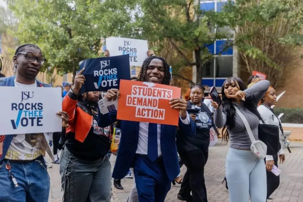 A group of Black students walk toward the polls, holding navy and white signs that say "JSU Rocks Thee Vote." In the center of the photo, a Black student wearing a navy blue suit holds a red Students Demand Action sign and smiles at the camera.