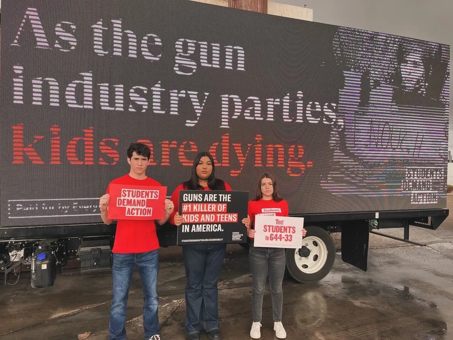 Three Students Demand Action volunteers stand in front of a billboard that reads "As the gun industry parties, kids are dying." They each hold signs. From left to right, the signs read: "Students Demand Action"; "Guns are the #1 killer of kids and teens in America"; "Text STUDENTS to 644-33."