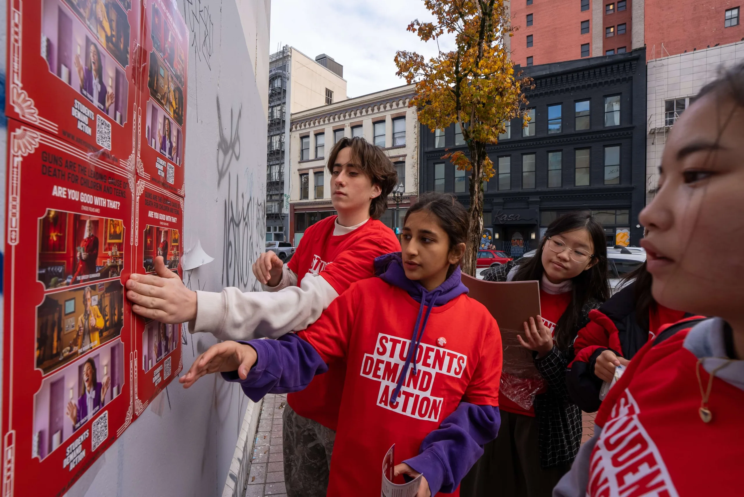 Four Students Demand Action volunteers hang posters in Portland, Oregon, during an in-person activation for the "Are You Good With That" industry accountability campaign. The posters are red and have black lettering at the top that says "Guns are the leading cause of death for children and teens." in white text beneath the black text, it says "Are you good with that?"