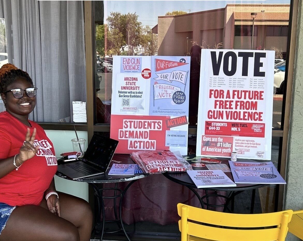 A young woman wears a red Students Demand Action t-shirt, denim shorts, and flip-flops. She is holding up a peace sign while sitting at a table with a laptop computer and two poster boards encouraging students on her campus to register to vote.