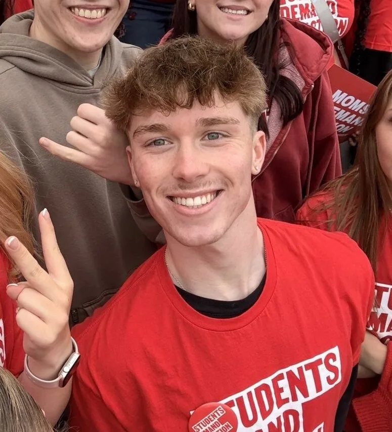 Brayden Burgess, one of the Students Demand Action Creators, poses for a selfie surrounded by several other students. He is wearing a red Students Demand Action t-shirt and a red Students Demand Action button.