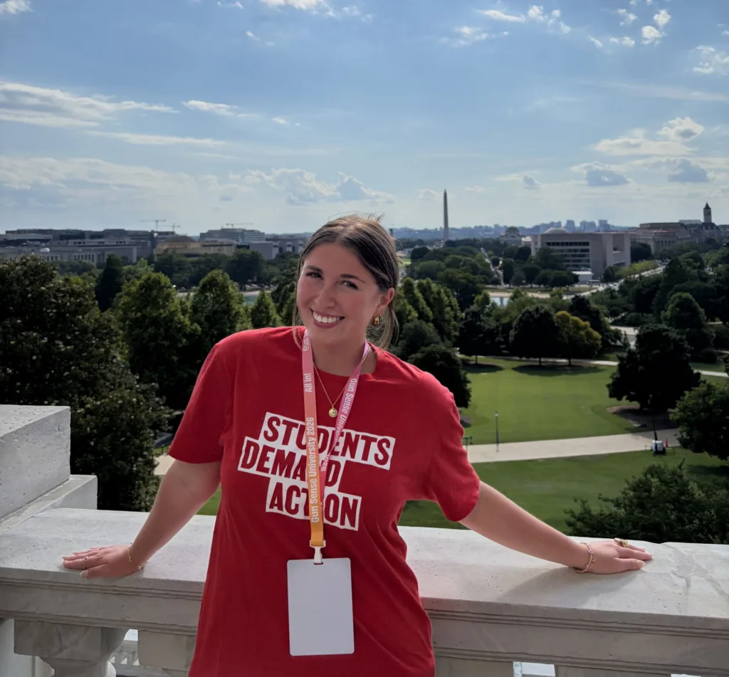 Cora, one of the Students Demand Action Creators, poses for a photo on a balcony with the Washington Monument in the background. She is wearing a red Students Demand Action t-shirt.