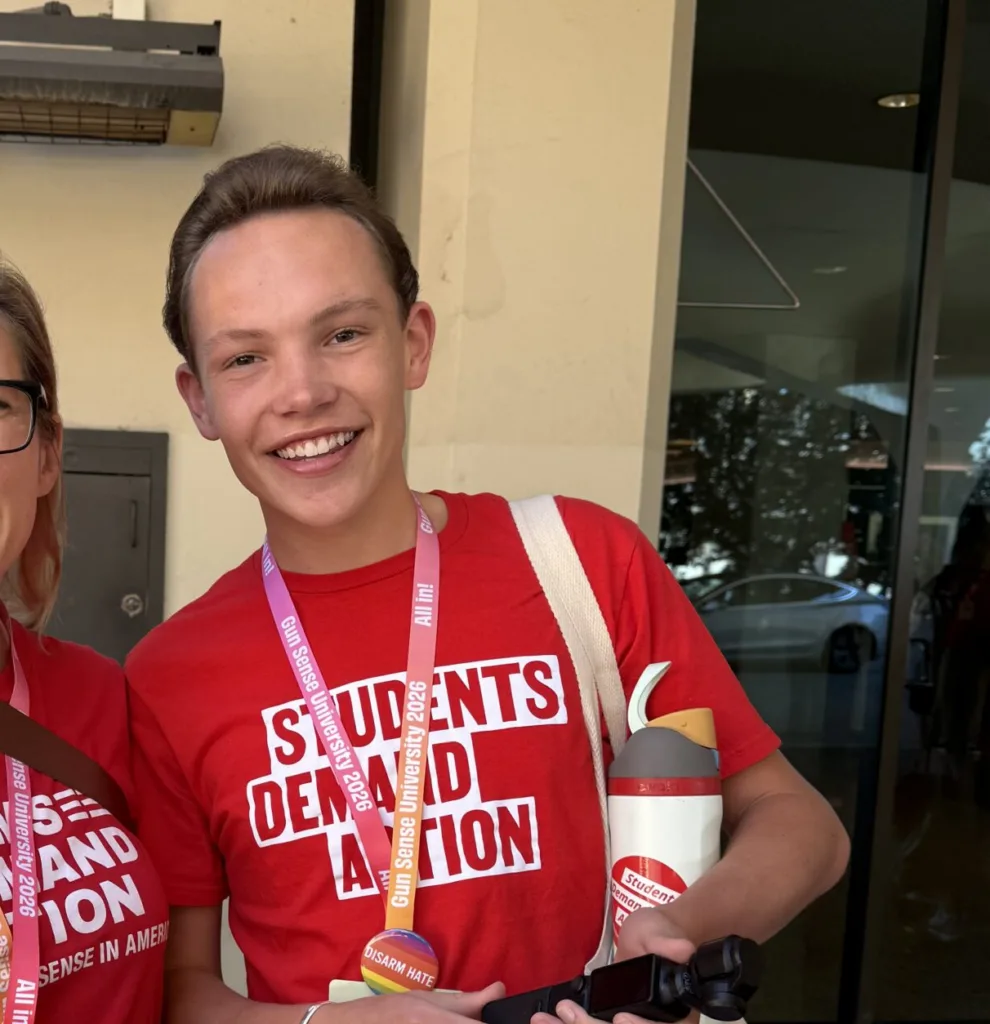 Charles, one of the Students Demand Action Creators, poses for a photo alongside a Moms Demand Action volunteer. Charles is wearing a red Students Demand Action t-shirt, white shorts, and is carrying a tripod, an Owala with a Students Demand Action sticker, and a canvas tote bag.