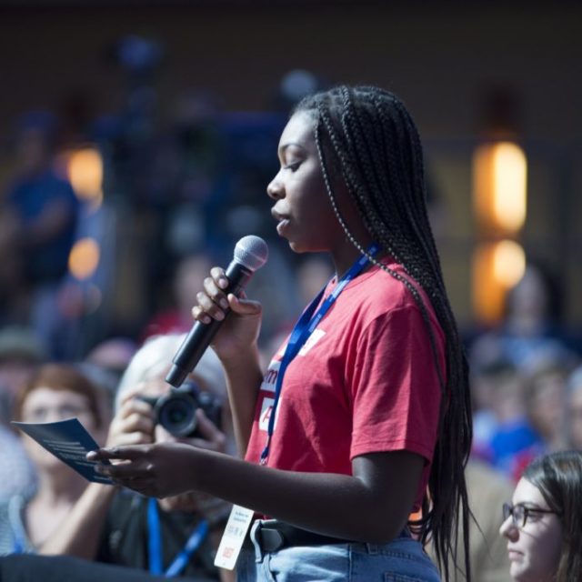 A Students Demand Action volunteer speaks in the microphone at the Presidential Gun Sense Forum. Behind them is a large crowd of other Moms Demand Action and Students Demand Action volunteers.