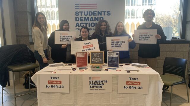 Megan Dombrowski and other members of Students Demand Action stand behind a SDA table holding signs that read Students Demand Action
